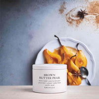 A white jar of KRINGLE Brown Butter Pear facial mask sits on a light wooden surface in front of a white plate of cooked pears with star anise and a spoon, against a textured blue-gray background.