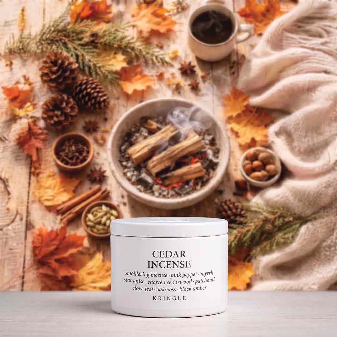 A white jar of KAINGLE Cedar Incense placed on a light surface, surrounded by autumn leaves, pinecones, cinnamon sticks, a cup of dark coffee, and a cream knit blanket in a cozy styled setting.