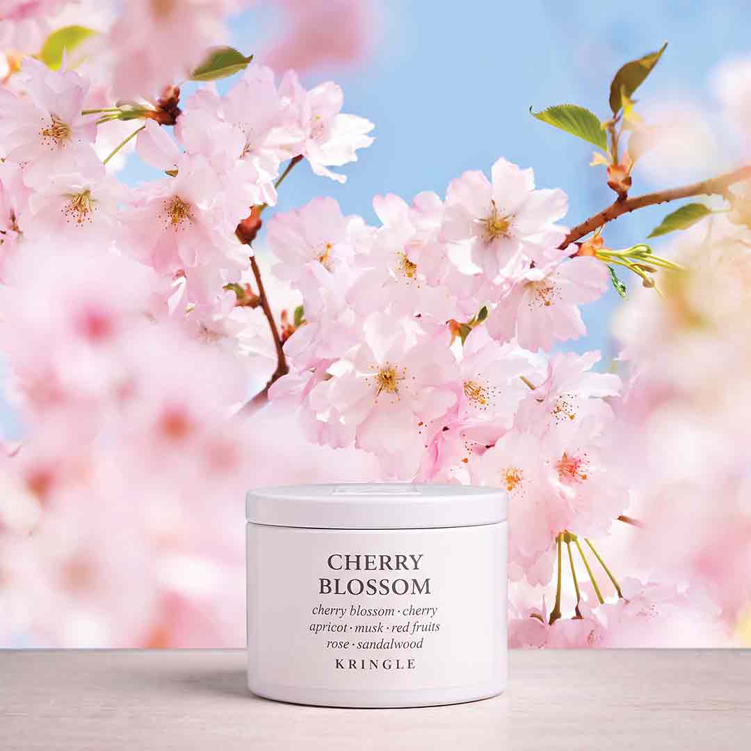 A white cosmetic jar labeled 'CHERRY BLOSSOM' by KINGLE sits on a light surface, surrounded by soft pink cherry blossoms and a blue sky background.