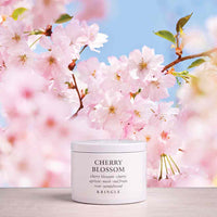 A white cosmetic jar labeled 'CHERRY BLOSSOM' by KINGLE sits on a light surface, surrounded by soft pink cherry blossoms and a blue sky background.