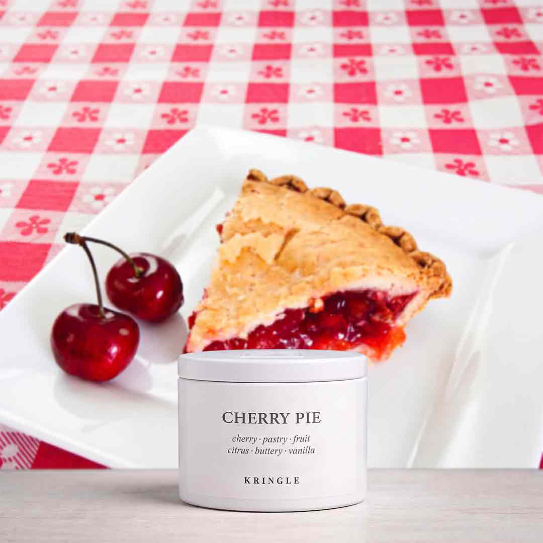 A white cylindrical tin labeled 'CHERRY PIE' and 'KRINGLE' sits in front of a slice of cherry pie on a white plate, with two red cherries and a red-and-white checkered tablecloth in the background.