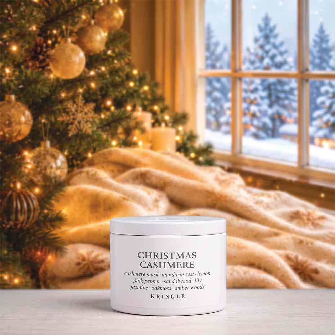 A white jar of KRINGLE Christmas Cashmere body wash sits on a tabletop in a cozy holiday setting, with a decorated Christmas tree, warm throw blanket, and window showing a snowy outdoor scene in the background.