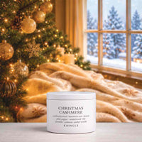 A white jar of KRINGLE Christmas Cashmere body wash sits on a tabletop in a cozy holiday setting, with a decorated Christmas tree, warm throw blanket, and window showing a snowy outdoor scene in the background.