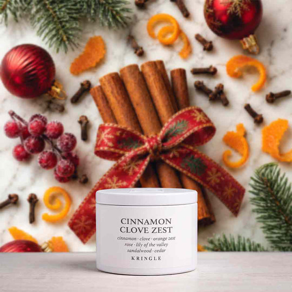 A white Kringle candle jar labeled 'Cinnamon Clove Zest' sits on a light surface, styled with cinnamon sticks tied in a red patterned ribbon, holiday ornaments, orange curls, cloves, pine branches, and frosted berries.