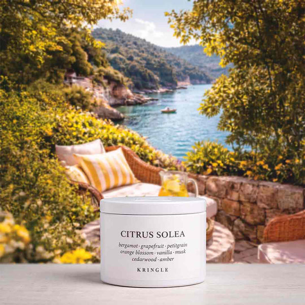 A white skincare jar labeled 'CITRUS SOLEA' by KRINGLE sits on a light stone surface in front of a scenic coastal patio backdrop with greenery, cushions, and blue water.