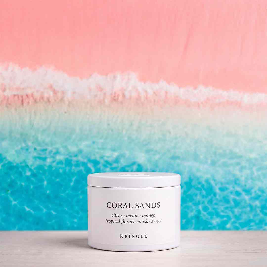 A white cylindrical jar of KRINGLE Coral Sands scented wax melt, placed on a light wooden surface against a vibrant beach-themed background with pink sky and turquoise ocean waves.