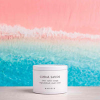 A white cylindrical jar of KRINGLE Coral Sands scented wax melt, placed on a light wooden surface against a vibrant beach-themed background with pink sky and turquoise ocean waves.
