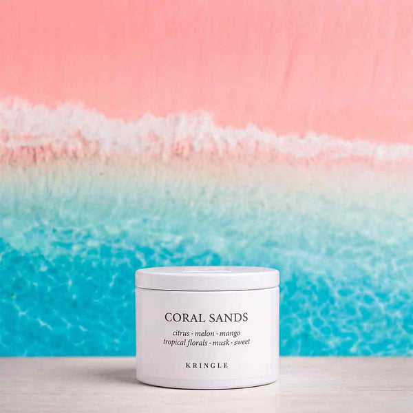A white cylindrical jar of KRINGLE Coral Sands scented wax melt, placed on a light wooden surface against a vibrant beach-themed background with pink sky and turquoise ocean waves.