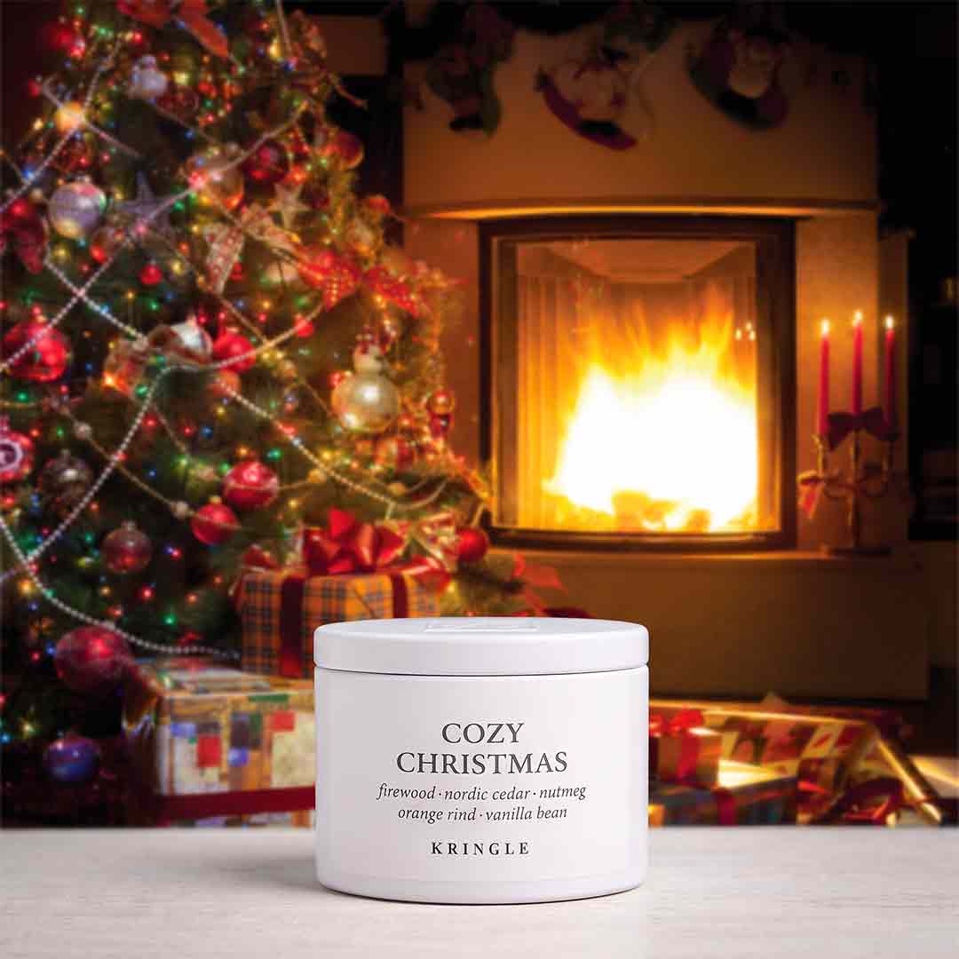 A white cylindrical jar of KRINGLE Cozy Christmas candle placed in front of a warm fireplace, with a decorated Christmas tree and wrapped gifts in the background.