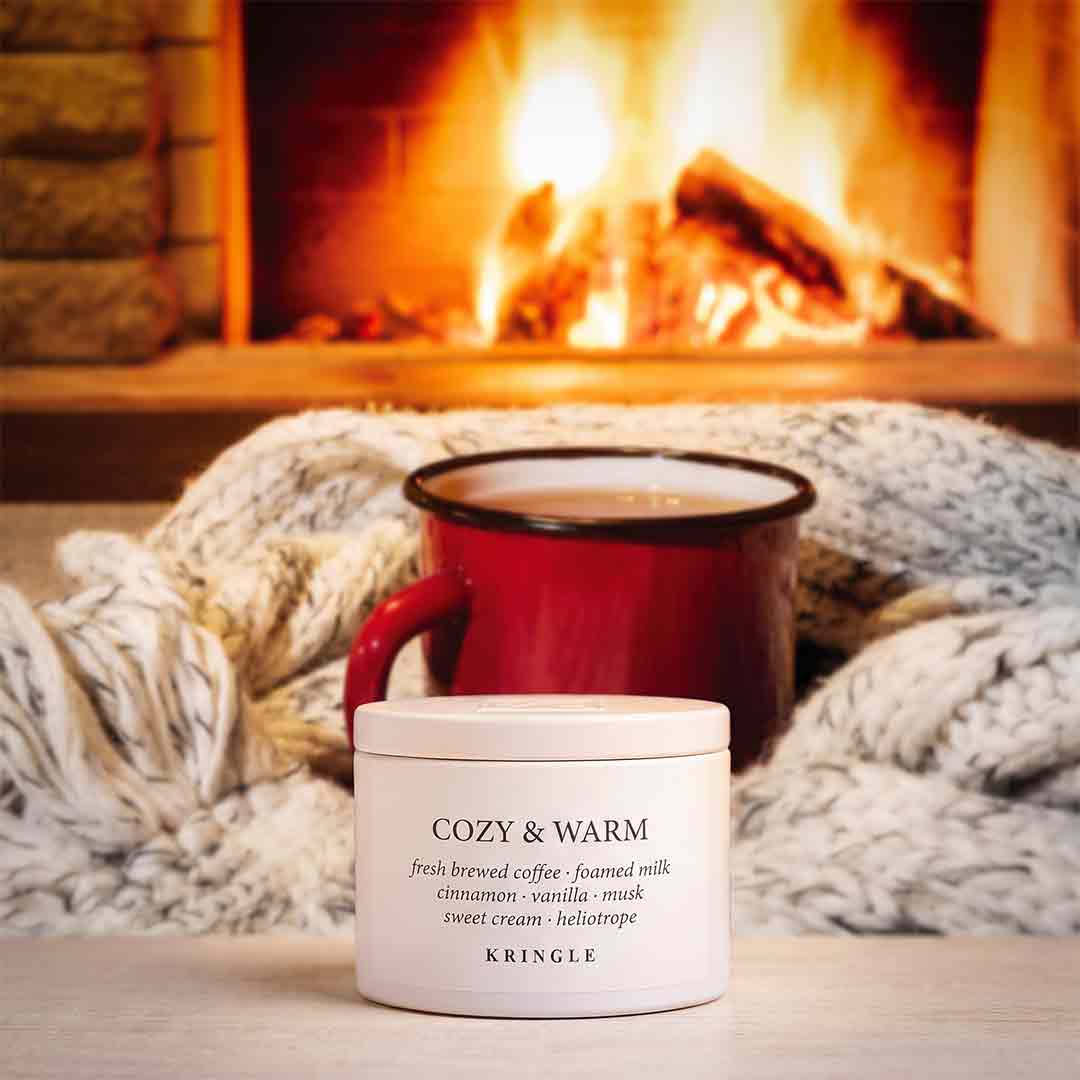 A white jar of KRINGLE Cozy & Warm scented candle sits in front of a red mug filled with coffee, wrapped in a chunky knit blanket, with a warm fireplace in the background.
