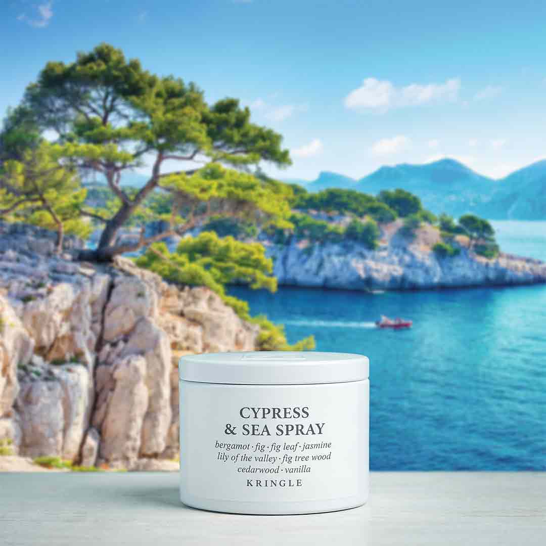 A white cylindrical jar of Kringle Cypress & Sea Spray placed on a light surface with a blurred coastal background showing blue water, rocky cliffs, green trees, and mountains.