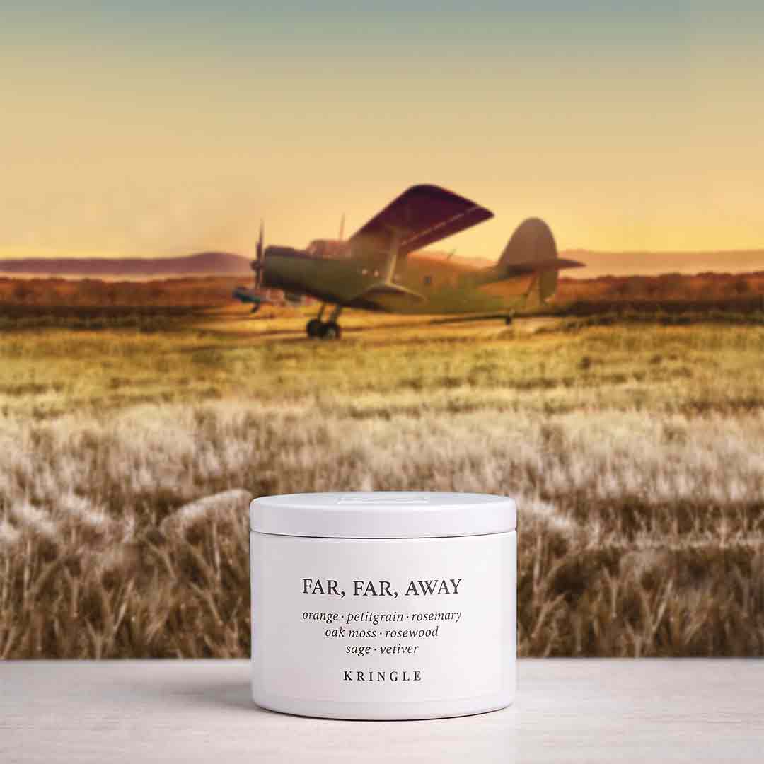 A white jar of KRINGLE 'Far, Far, Away' body butter sits on a light surface in the foreground, with a blurred background of a vintage airplane landing in a grassy field at sunset.