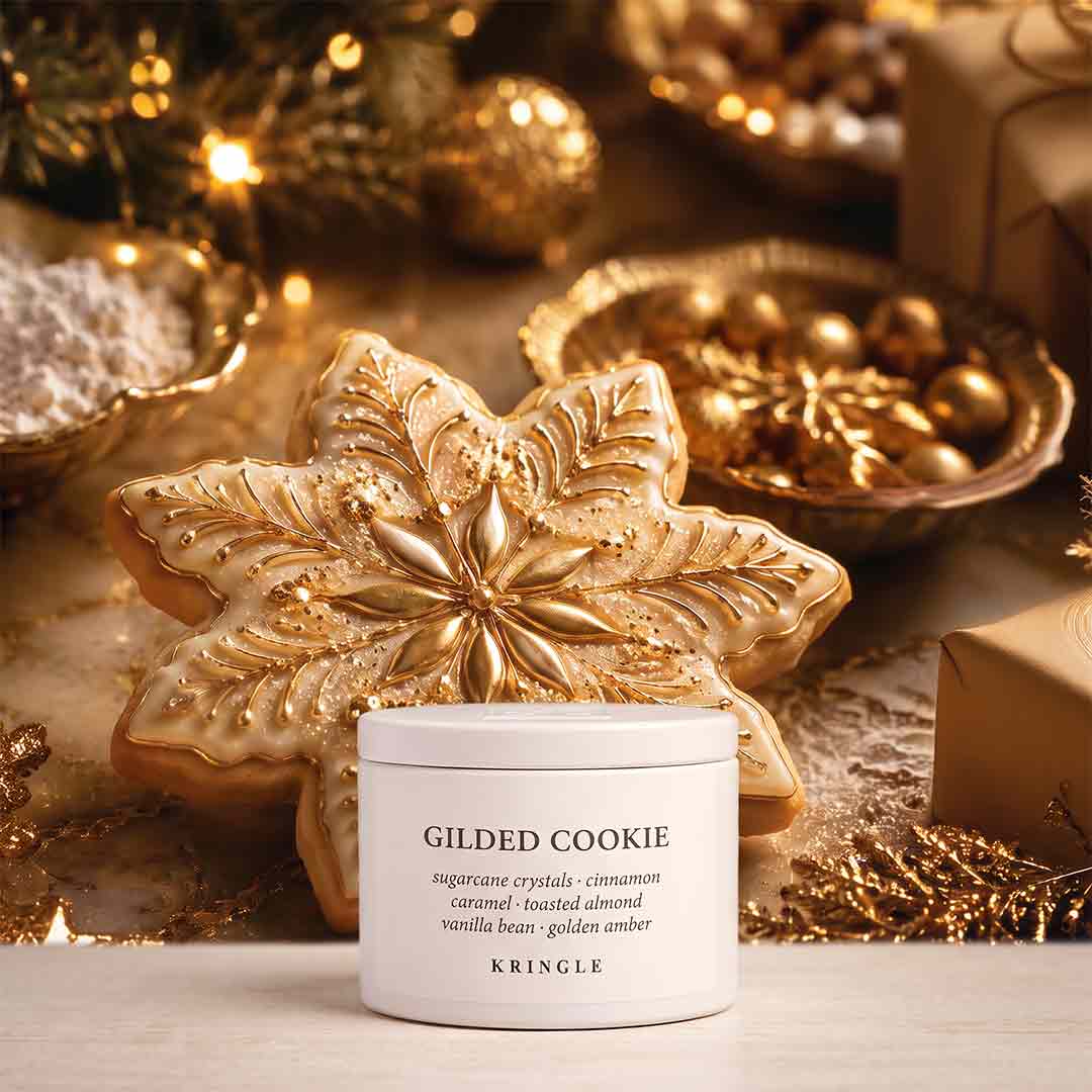 A white jar of KRINGLE Gilded Cookie scented candle placed in front of a large decorated gilded cookie, with warm holiday ornaments, lights, and wrapped gifts in the background.