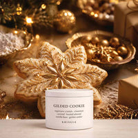 A white jar of KRINGLE Gilded Cookie scented candle placed in front of a large decorated gilded cookie, with warm holiday ornaments, lights, and wrapped gifts in the background.