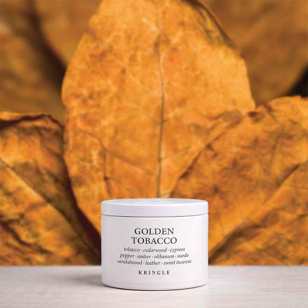 A white cylindrical jar of Kringle Golden Tobacco incense placed on a light surface, with large dried golden-brown leaves forming a warm, blurred background.