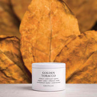 A white cylindrical jar of Kringle Golden Tobacco incense placed on a light surface, with large dried golden-brown leaves forming a warm, blurred background.