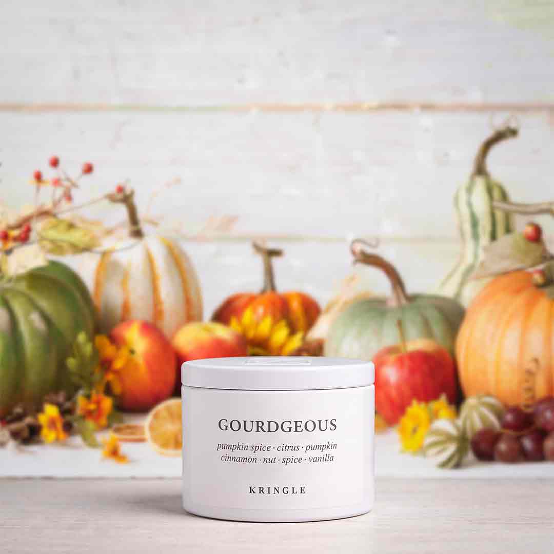 A white jar of KRINGLE GOURDEOUS pumpkin spice candle sits on a light wooden surface, styled with autumn pumpkins, apples, and small yellow flowers in the background.