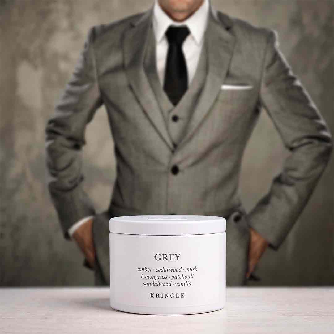 A white jar of KRINGLE Grey Lemongrass Patchouli Sandalwood Vanilla body butter is placed on a light surface, with a blurred man in a grey suit standing in the background.