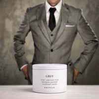 A white jar of KRINGLE Grey Lemongrass Patchouli Sandalwood Vanilla body butter is placed on a light surface, with a blurred man in a grey suit standing in the background.
