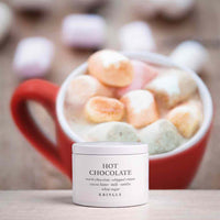 A white jar of KINGLE Hot Chocolate whipped cream sits in the foreground, with a blurred red mug filled with marshmallows and hot chocolate in the background.