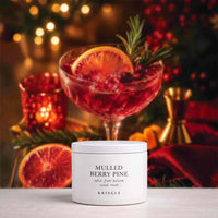 A white cylindrical candle tin labeled 'MULLED BERRY PINE' by KRINGLE sits on a light surface in front of a festive holiday scene with a red berry cocktail, orange slices, greenery, and warm blurred lights.