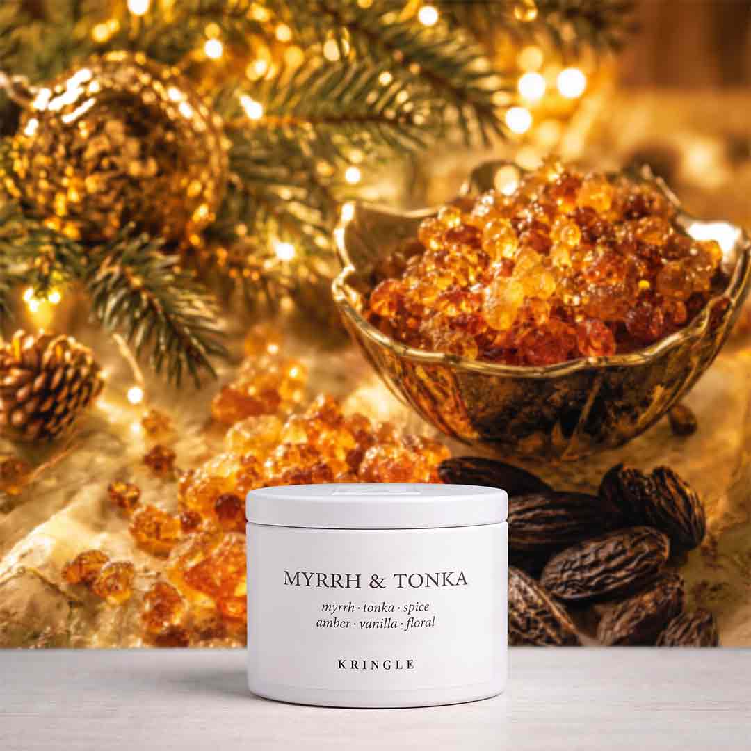A white cylindrical jar labeled 'MYRRH & TONKA' by KRINGLE sits on a light surface, surrounded by amber resin pieces, dark brown spices, pine branches, and warm golden fairy lights in the background.