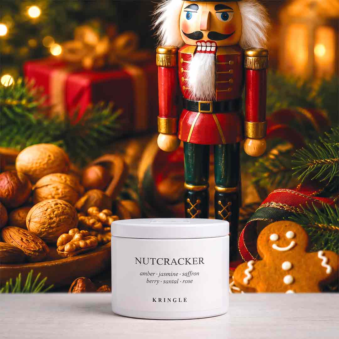A white cylindrical candle tin labeled 'NUTCRACKER' by KRINGLE sits on a light surface in a festive holiday setting with a wooden nutcracker, nuts, pine branches, wrapped gifts, and a gingerbread cookie in the background.
