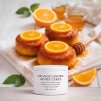 A white jar of KRINGLE Orange Ginger Honey Cakes fragrance centered in the foreground, with three orange-topped baked cakes, sliced oranges, honey, and a wooden dipper in the background on a light surface.