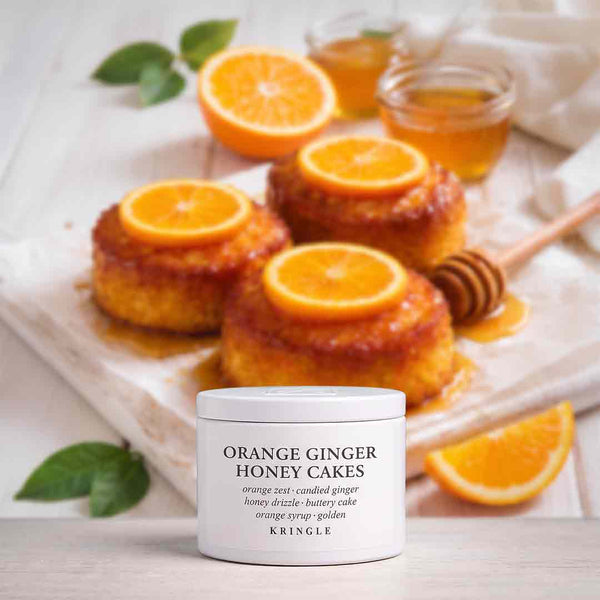 A white jar of KRINGLE Orange Ginger Honey Cakes fragrance centered in the foreground, with three orange-topped baked cakes, sliced oranges, honey, and a wooden dipper in the background on a light surface.