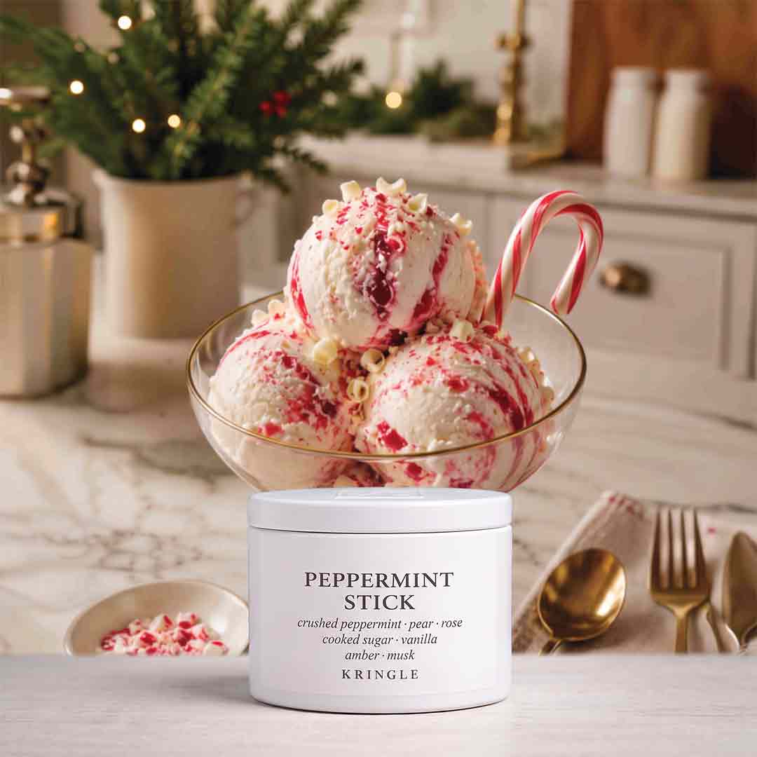 A white jar of KRINGLE Peppermint Stick scented candle sits in front of a glass bowl filled with vanilla ice cream topped with crushed candy cane pieces and a candy cane stick, on a marble surface in a softly blurred holiday kitchen setting.