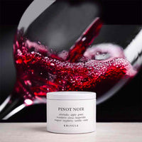A white jar of KRINGLE Pinot Noir body butter sits on a light surface in front of a blurred glass of red wine.