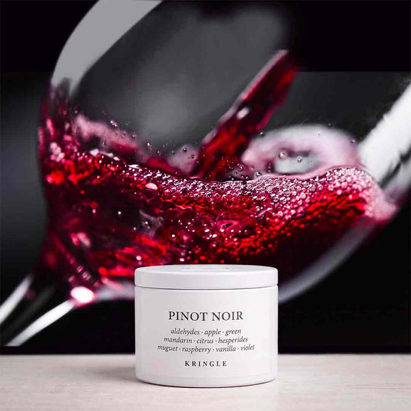 A white jar of KRINGLE Pinot Noir body butter sits on a light surface in front of a blurred glass of red wine.