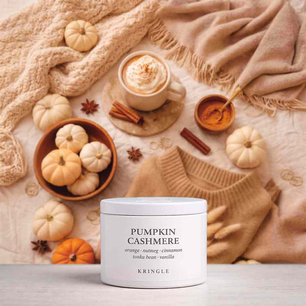 A white jar candle labeled 'PUMPKIN CASHMERE' by KRINGLE sits on a light surface, styled with a beige sweater, knit blanket, small pumpkins, cinnamon sticks, a cup of hot cocoa, and star anise.