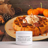A white jar of KRINGLE Pumpkin Waffles scented candle sits on a light surface in the foreground, with a plate of pumpkin waffles topped with whipped cream and pecans blurred in the background against a rustic wood setting with pumpkins.