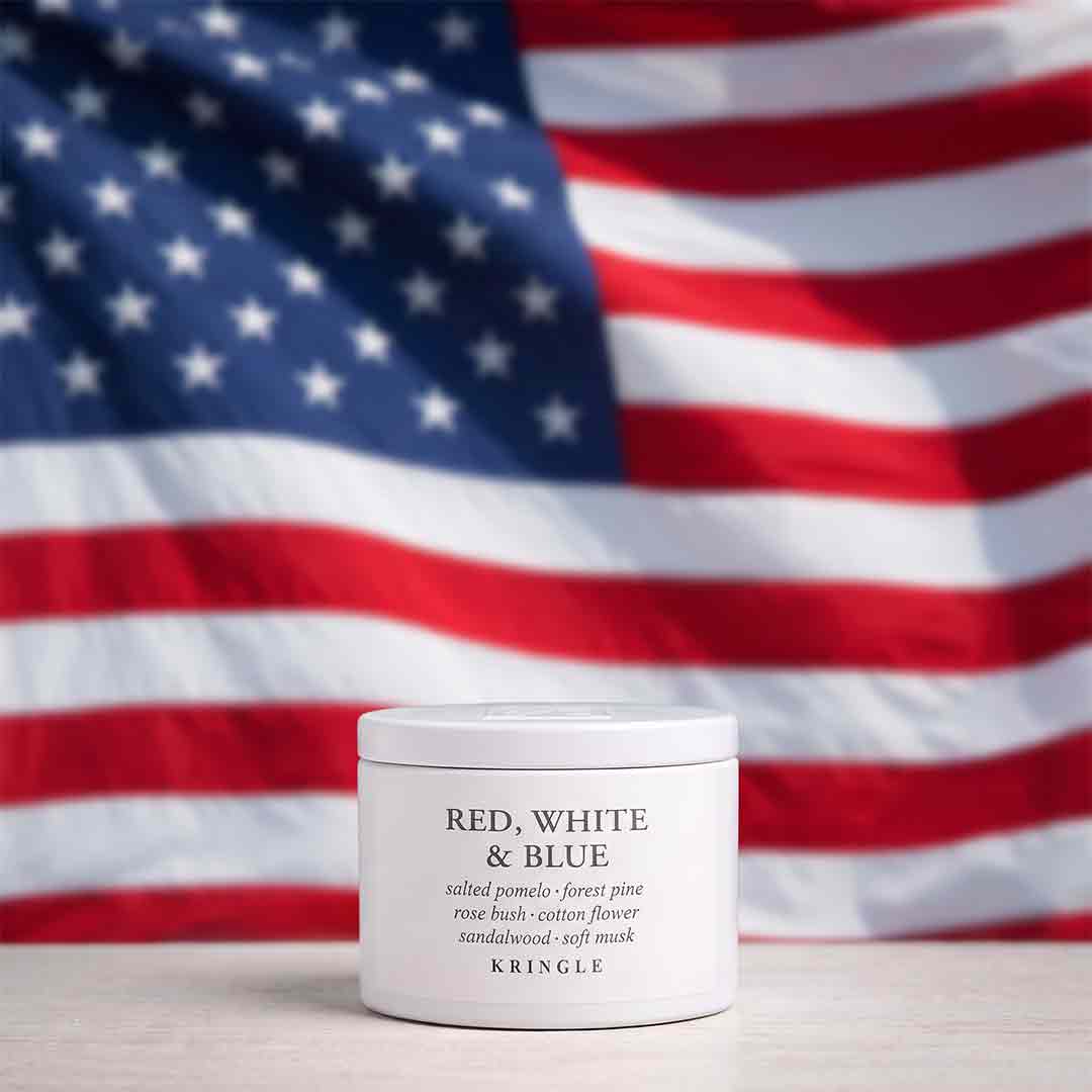 A white jar of KRINGLE 'Red, White & Blue' salined pomade placed on a light surface with a blurred American flag in the background.