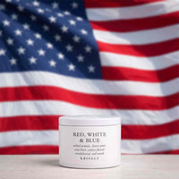 A white jar of KRINGLE 'Red, White & Blue' salined pomade placed on a light surface with a blurred American flag in the background.