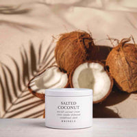 A white jar of KRINGLE Salted Coconut sea salt coconut-cream clay-matza drywood and sandalwood mask sits on a light surface, styled with whole and halved coconuts and palm leaf shadows against a beige background.