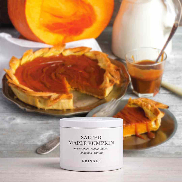 A white jar of KRINGLE Salted Maple Pumpkin spread placed in the foreground on a light gray surface, with a sliced pumpkin pie, a slice on a plate, a glass of syrup, and a pumpkin in the background.