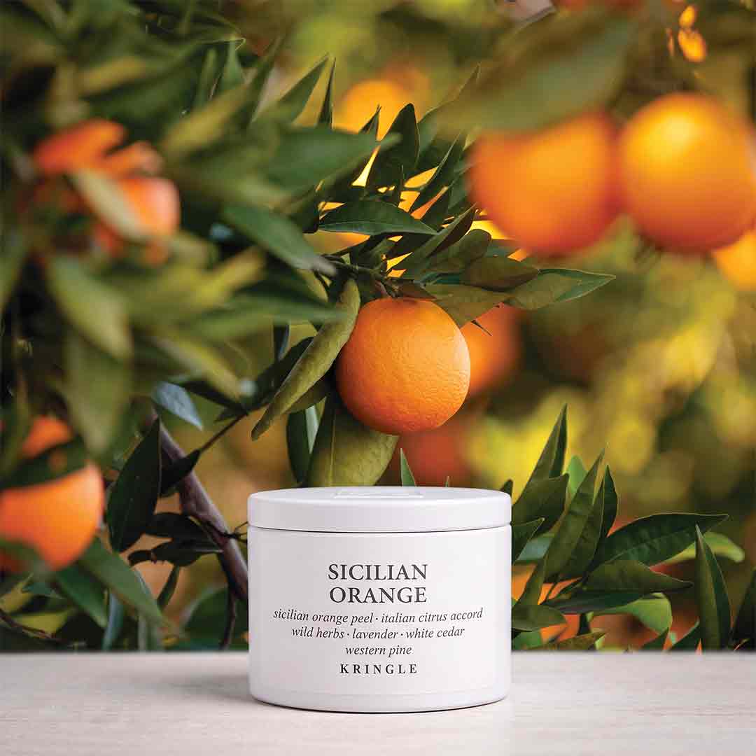 A white jar of KRINGLE Sicilian Orange peel and citrus accordo body scrub sits on a light surface in front of a blurred orange tree with ripe oranges and green leaves.