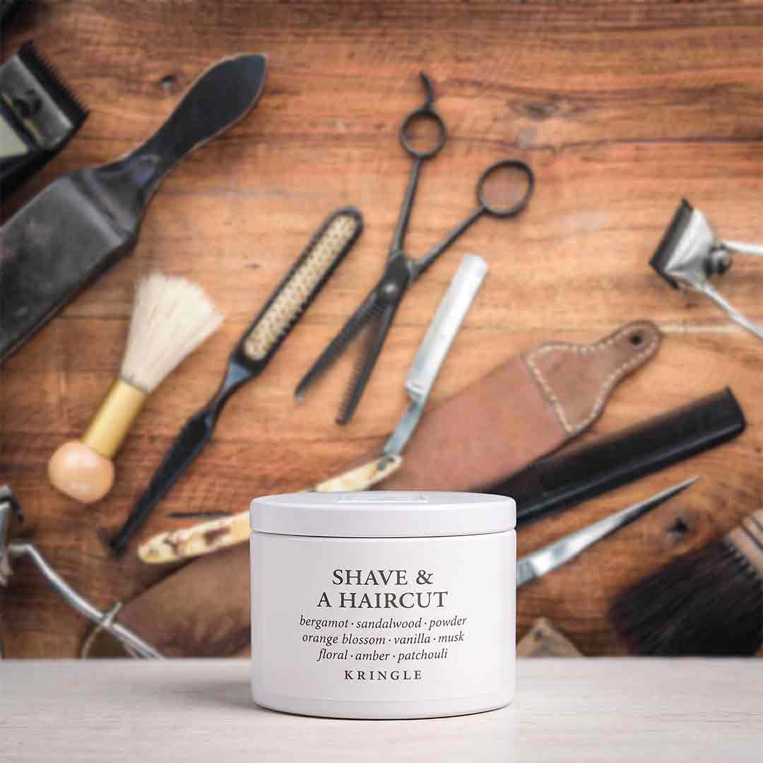 A white jar of Kringle Shave & A Haircut grooming product sits on a light surface, surrounded by barber tools and brushes on a rustic wooden background.