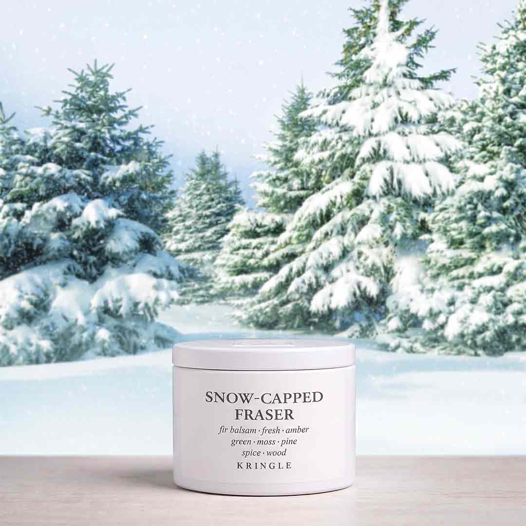 A white cylindrical tin of KRINGLE Snow-Capped Fraser balmy body butter sits on a light wooden surface with a snowy pine forest background.