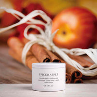 A white jar of KRINGLE Spiced Apple mask placed on a wooden surface, with cinnamon sticks and red apples in the background.