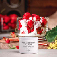 A white jar of KRINGLE Strawberry & Cream whipped cream sits on a wooden surface in front of a glass dessert filled with fresh strawberries and white cream, with strawberries and leaves in a softly blurred background.