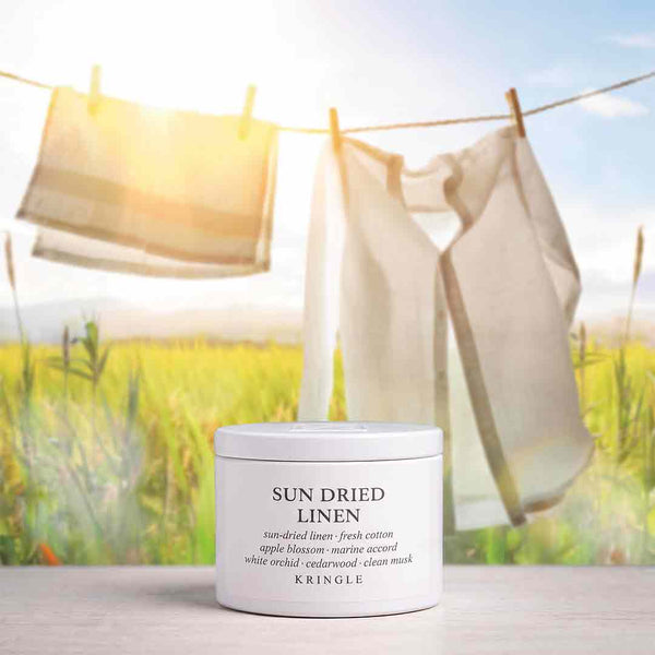 A white cylindrical jar of KRINGLE Sun Dried Linen mask placed on a light surface, with a blurred outdoor scene behind it showing hanging beige towels or sheets in front of a bright sky and field.