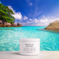 A white jar of KRINGLE Tropical Waters body butter or scrub stands on a light surface in front of a tropical beach backdrop with turquoise water, blue sky, and green trees. The label reads 'TROPICAL WATERS' with scent notes and the brand KRINGLE visible.