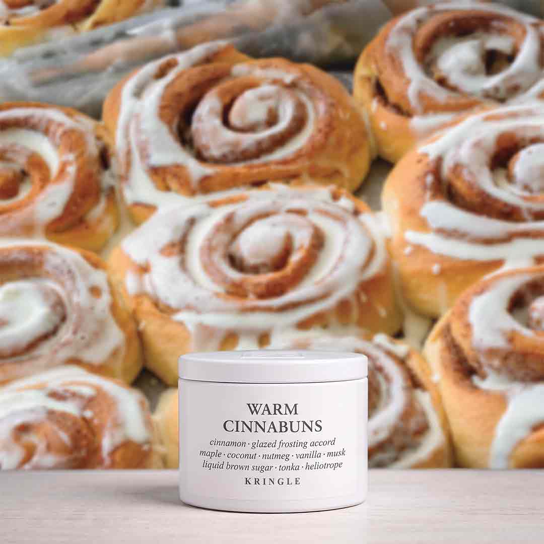 A white jar of KRINGLE Warm Cinnabuns cosmetic product placed in front of a blurred background of cinnamon rolls with white icing.