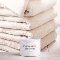 A white jar of KRINGLE Warm Cotton mask placed in front of a stack of folded cream-colored towels on a light wooden surface.