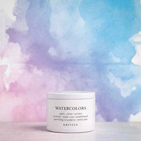 A white jar of KRINGLE Watercolors face mask, placed on a light surface against a colorful watercolor background of blue, purple, and pink.