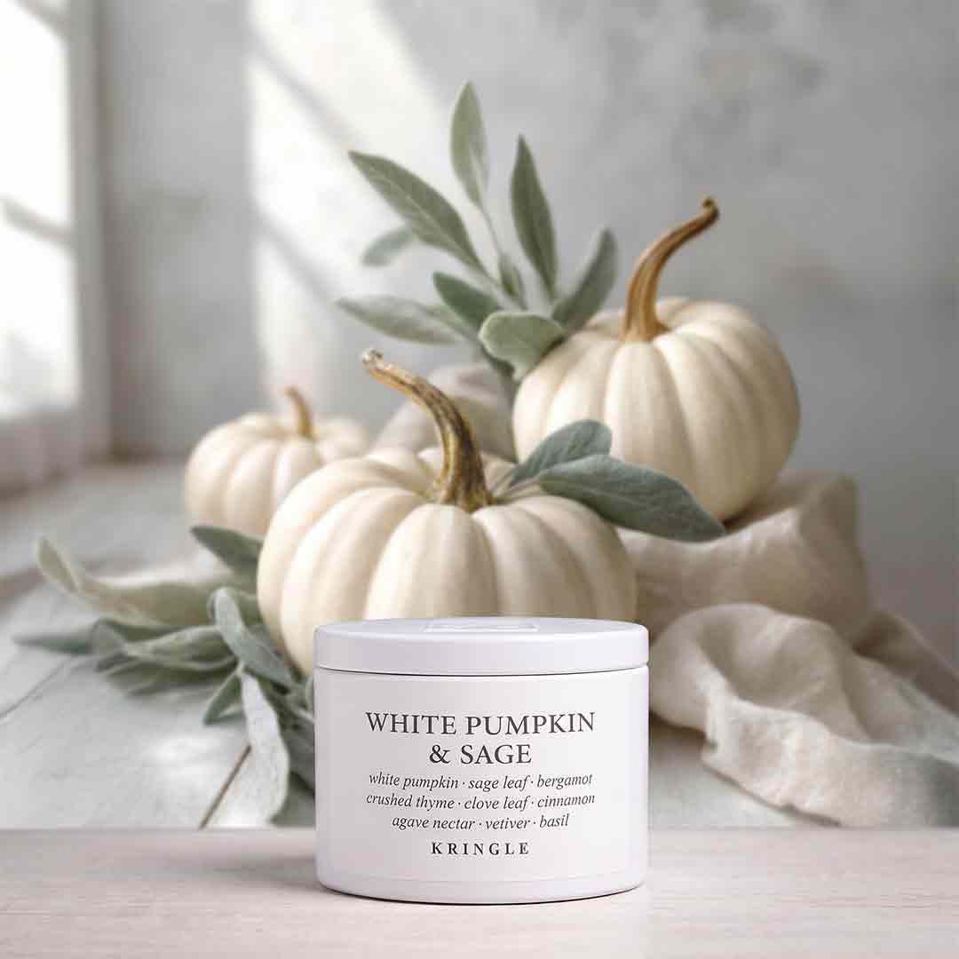 A white jar candle labeled 'WHITE PUMPKIN & SAGE' by KRINGLE sits on a light surface, styled with cream pumpkins, sage leaves, and a draped fabric in a soft, bright setting.