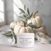 A white jar candle labeled 'WHITE PUMPKIN & SAGE' by KRINGLE sits on a light surface, styled with cream pumpkins, sage leaves, and a draped fabric in a soft, bright setting.